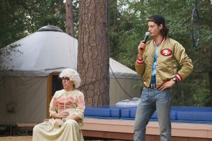 A person in a long dress, wig, and sunglasses sits holding a fan, while another person in a 49ers jacket and backwards cap stands nearby, speaking into a microphone, outdoors near a yurt and trampoline.