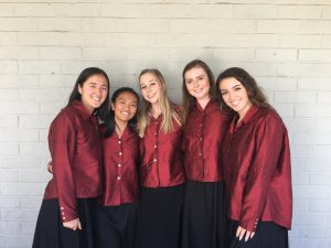 Five young women stand side by side, smiling at the camera. They are wearing matching maroon blouses and black skirts, posing in front of a light gray brick wall.