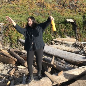 A person with shoulder-length dark hair, wearing a plaid shirt and leggings, stands on driftwood at the beach, holding a yellow bottle and smiling with one arm playfully raised. Green plants cover the slope behind them.