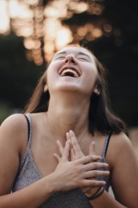 A person with long dark hair laughs joyfully, head tilted back and hands clasped to their chest, wearing a gray sleeveless top. The background is softly blurred with warm light.