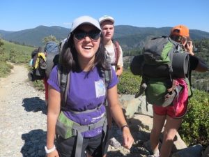 Three young adults with backpacks hike on a rocky trail in the mountains under a clear blue sky. The woman in front is smiling, wearing sunglasses and a cap. The group is surrounded by greenery and distant hills.