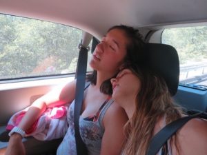 Two young women are sitting in the backseat of a car, both wearing seat belts and sleeping. One woman is resting her head on the others shoulder. Sunlight streams through the window beside them.