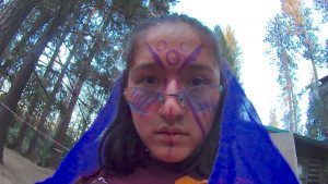 A person with a blue veil, face painted with purple geometric patterns, and wearing glasses, looks directly at the camera. Tall trees and a building are visible in the outdoor background.