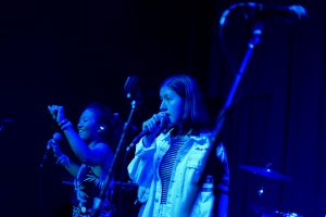 Two young women perform on stage under blue lighting; one sings into a microphone, while the other smiles and raises her arm. Musical equipment and microphones are visible in the background.