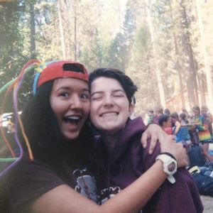 Two smiling teens hugging outdoors, wearing maroon sweatshirts. One has a red cap with colorful pipe cleaners. There are trees and other people in the background, suggesting a camp or outdoor event.