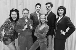 Six young adults pose together in front of a curtain, dressed in a variety of outfits including business suits, athletic wear, and casual clothing, suggesting a theatrical or themed event. They all look confidently at the camera.