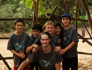 Six boys wearing matching camp T-shirts smile and pose together outdoors, standing in front of a rustic wooden fence surrounded by trees. One crouches in front while five stand closely behind him.