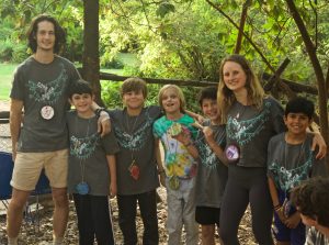 A group of six kids and two adults stand outdoors, smiling and posing together. They wear matching gray shirts with lanyards and medallions. Trees and greenery fill the background, suggesting a camp or nature setting.
