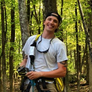 A smiling person wearing a white t-shirt, harness, and cap stands in a sunlit forest holding climbing gear.