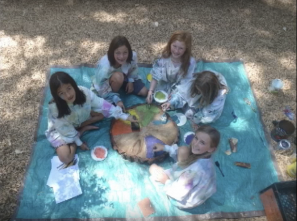 Five children sit on a teal mat outdoors, wearing paint-splattered shirts. They smile up at the camera while collaboratively painting a large, circular wooden slab with colorful designs. Art supplies are scattered nearby.