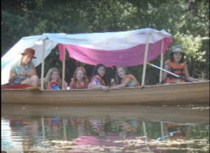 Six children wearing life jackets and colorful hats sit in a wooden canoe covered by a pink and white tarp, floating on a calm river with trees in the background.