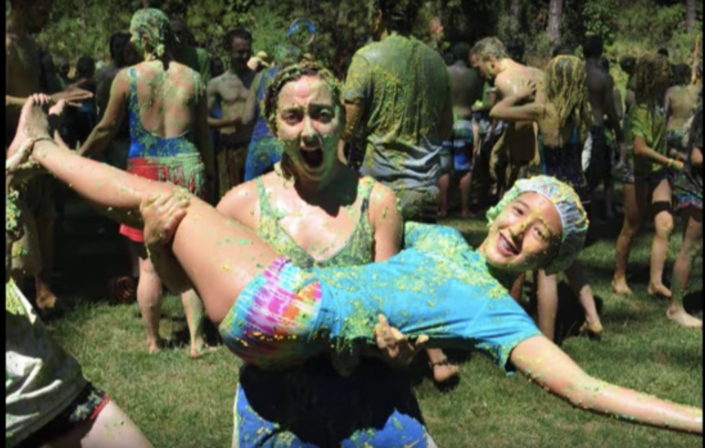 Two people covered in green slime smile and pose outdoors; one is holding the other horizontally. Other people in swimsuits and shorts, also covered in slime, are gathered in the background on grass.
