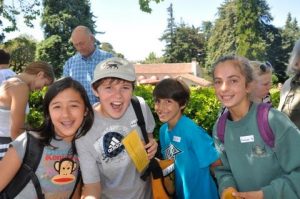 Four smiling children with backpacks stand outdoors in front of greenery on a sunny day. They appear excited and are holding papers. Several adults are visible in the background.