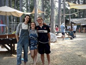 Three young adults stand smiling with arms around each other at an outdoor camp, surrounded by tall trees, sunshades, and other people in the background.