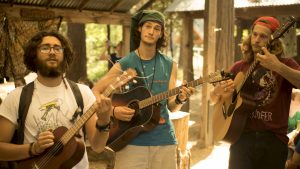 Three young men play guitars and a ukulele outdoors under a wooden shelter. They have long hair, beards, and casual clothing, with two wearing hats or bandanas. Sunlight filters through trees in the background.
