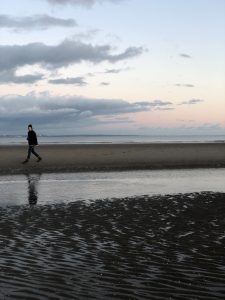 A person walks alone on a sandy beach near the water’s edge at dusk, with clouds and soft pastel colors in the sky reflecting the calm, tranquil mood of the scene.