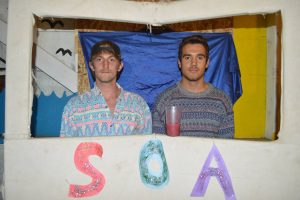 Two young men stand side by side in a homemade booth with colorful letters “S,” “O,” and “A” on the front. One man holds a drink; both wear patterned shirts. The background is decorated with blue and yellow fabric.