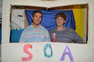 Two young men stand side by side behind a white frame decorated with glittery letters S, O, and A. One holds a drink, and both wear patterned shirts. The colorful backdrop features waves and a blue cloth.