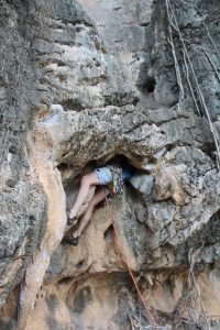 A rock climber wearing climbing gear squeezes through a narrow opening in a rugged, rocky cliff, with ropes and carabiners attached to their harness. Only the climbers lower body and legs are visible.