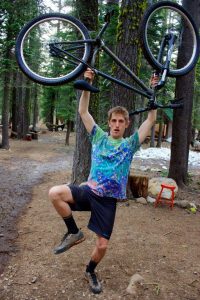 A man stands in a forest, holding a bicycle above his head with both hands while balancing on one leg. He wears a colorful t-shirt, dark shorts, and socks with shoes. Trees and a red stool are in the background.