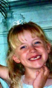 A young girl with blond hair wearing a small tiara smiles widely, showing missing teeth. She is wearing a light-colored dress and standing in front of a light background.