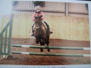 A child wearing a helmet and red jacket rides a dark horse, jumping over green and white poles in an indoor riding arena.