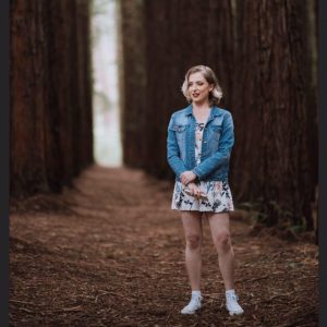 A person in a blue denim jacket, floral dress, and white sneakers stands on a path surrounded by tall trees in a forest, looking slightly to the side and smiling.