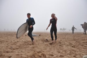 Two people in wetsuits walk on a sandy beach, one carrying a surfboard. The background is foggy, with faint figures and camels visible in the distance.
