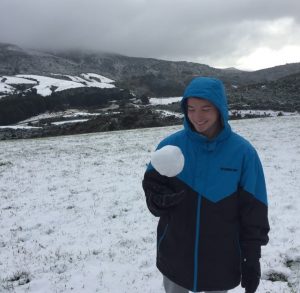 A person in a blue and black winter jacket stands on a snowy field, smiling while holding a large snowball. Mountains and cloudy skies are visible in the background.
