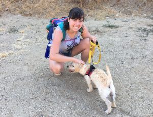 A woman with a backpack smiles while kneeling to pet a small, light-colored dog wearing a harness and leash on a dry, dirt path with dried grass in the background.