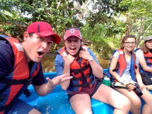 Four young people wearing life vests sit in a blue boat on a river, surrounded by lush green trees. Two in front are excited and smiling at the camera, while the two in back are more relaxed.