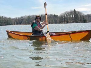 A woman in a green shirt and life jacket paddles a wooden canoe on a lake, smiling and splashing water, with trees and a clear sky in the background.