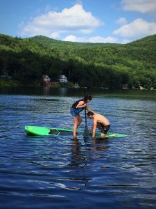Two people on a calm lake, one standing on a paddleboard helping the other, who is kneeling on a board. Green hills, trees, and houses are visible in the background under a blue sky with clouds.