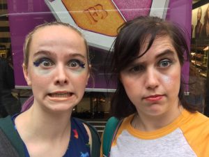 Two young women with dramatic, colorful makeup make silly faces in front of a makeup store window display featuring a large compact. Both have shimmery eye makeup and exaggerated expressions.