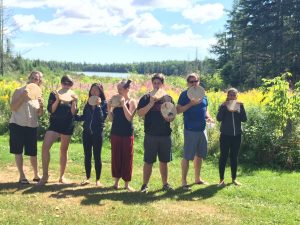Seven people stand outdoors in a grassy area by a lake, holding round, flat drum-like objects and posing playfully. Trees, wildflowers, and a blue sky with clouds are visible in the background.
