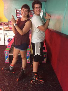 Two people wearing rollerblades stand back-to-back indoors, smiling and posing with finger guns in front of a brightly-colored arcade game. The setting has teal walls and dark patterned carpet.