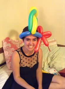 A smiling person in a black dress with sheer polka-dot top sits on a bed, wearing a colorful balloon hat made of yellow, blue, red, and green balloons twisted together.