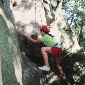 A child wearing a red helmet, green shirt, and red shorts climbs a rock wall outdoors, secured with a harness and rope, surrounded by trees and sunlight.