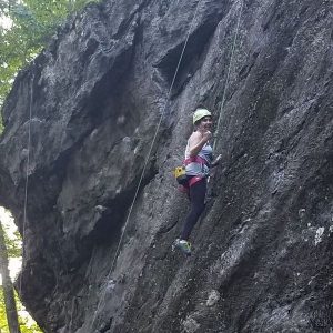 A person wearing a helmet and climbing gear ascends a steep, rocky cliff face, gripping the rock and attached to safety ropes. Trees are visible at the top left of the image.