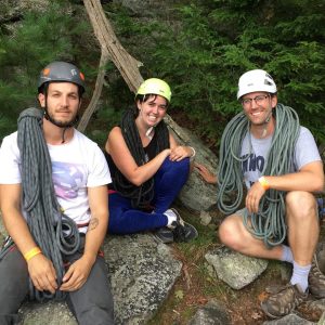 Three people in helmets and climbing gear sit on rocks outdoors, smiling. Coiled climbing ropes hang around their shoulders. Trees and greenery are in the background.