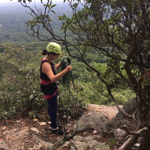 A person wearing a helmet and harness coils a climbing rope near the edge of a rocky cliff, surrounded by trees, with a forested valley visible in the background.
