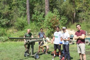 Six boys stand outdoors near a homemade green catapult or launcher on a grassy area, surrounded by trees and bushes. Some are holding projectiles and smiling at the camera. The weather appears sunny.