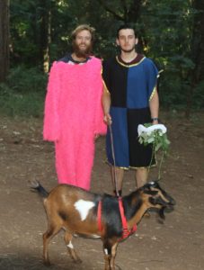 Two men stand in a wooded area; one wears a bright pink furry suit, the other a black and blue tunic, holding a leash attached to a brown goat. The man in the tunic also holds a leafy branch.