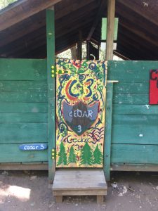 A green wooden cabin with a colorful, hand-painted door that says “CEDAR 3” and features trees and abstract designs. A small wooden step leads up to the door. A blue sign on the left also reads “cedar 3.”.