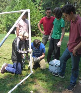 A group of teens outdoors assembles a white PVC pipe structure. One kneels to attach a pipe while others watch or assist. Trees and grass fill the background, and a box sits on the ground nearby.