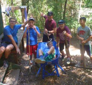 Six boys are outdoors in a wooded area, posing playfully. One boy sits in a wheelbarrow while the others stand around him, holding rakes and sticks, smiling and making animated gestures. It appears to be a camp setting.