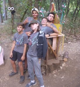 Five kids wearing matching camp T-shirts pose together in a wooded area, some making playful gestures and faces, near a wooden structure decorated with leaves and flowers.