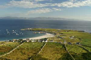 A coastal village with scattered houses, green fields separated by stone walls, a sandy beach, and boats anchored in the calm blue sea, under a partly cloudy sky with distant hills across the water.