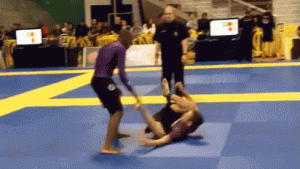 Two athletes in purple rash guards compete in a grappling match on blue mats, as one tries to pass the others guard while a referee and crowd watch in the background.
