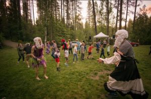 People dance and play on a grassy field surrounded by trees. Two adults in wigs and costumes face each other while a group of children and adults gather and watch or join in the fun. A tent and parked car are in the background.